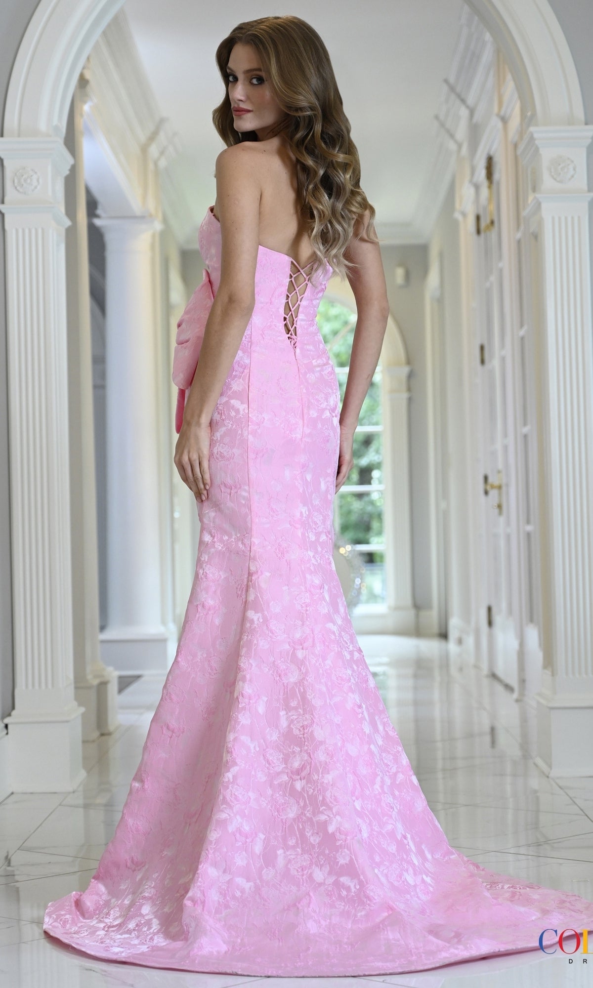 A woman wears a floor-length pink floral jacquard mermaid Long Prom Dress: Colors Dress 4065 with a strapless neckline, lace-up back, and a side bow while standing in a hallway.